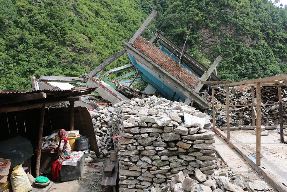 A fallen building at the base of a mountain, and a person sitting under a sloping roof alongside a large pile of bricks.