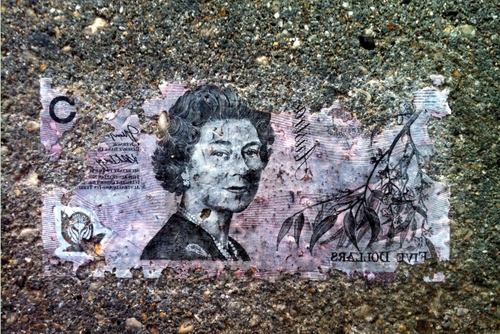 Reverse image of colorful Australian five dollar currency note ground into rocky pavement with image of Queen Elizabeth the II looking over her right shoulder toward viewer.