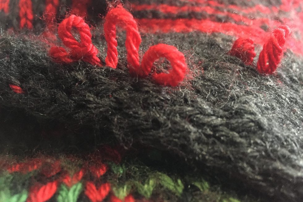 Close-up photograph of a piece of knitting. The photograph focuses on unraveling red loops of yarn poking out of black fabric. Different patterns of red, green, and black knitting appear in the foreground and background of the photograph.